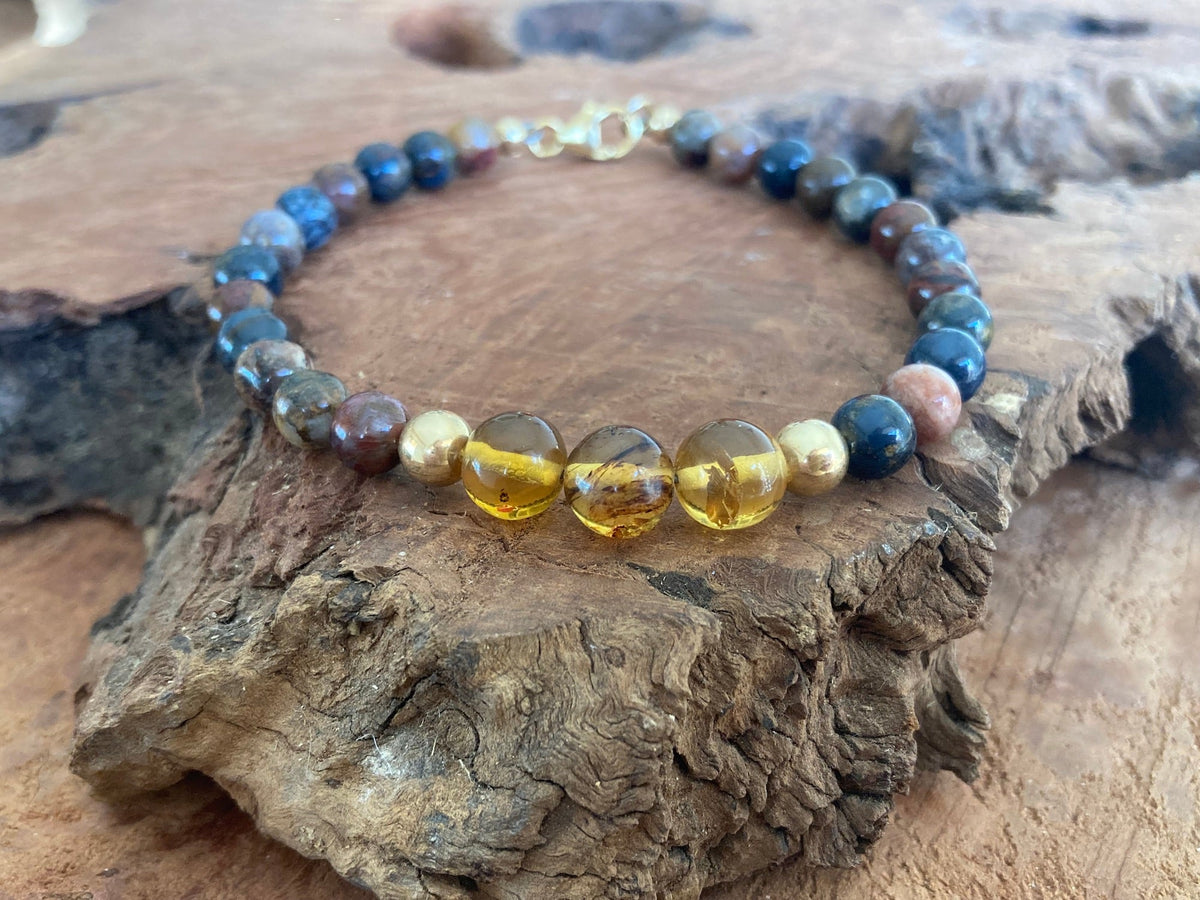 The Catalyst bracelet's honey-coloured amber beads in front, resting on a driftwood slab