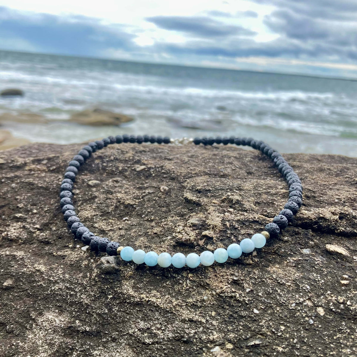 Courage men's necklace: pale blue aquamarine with dark lava stone beads, laid on rock by the sea