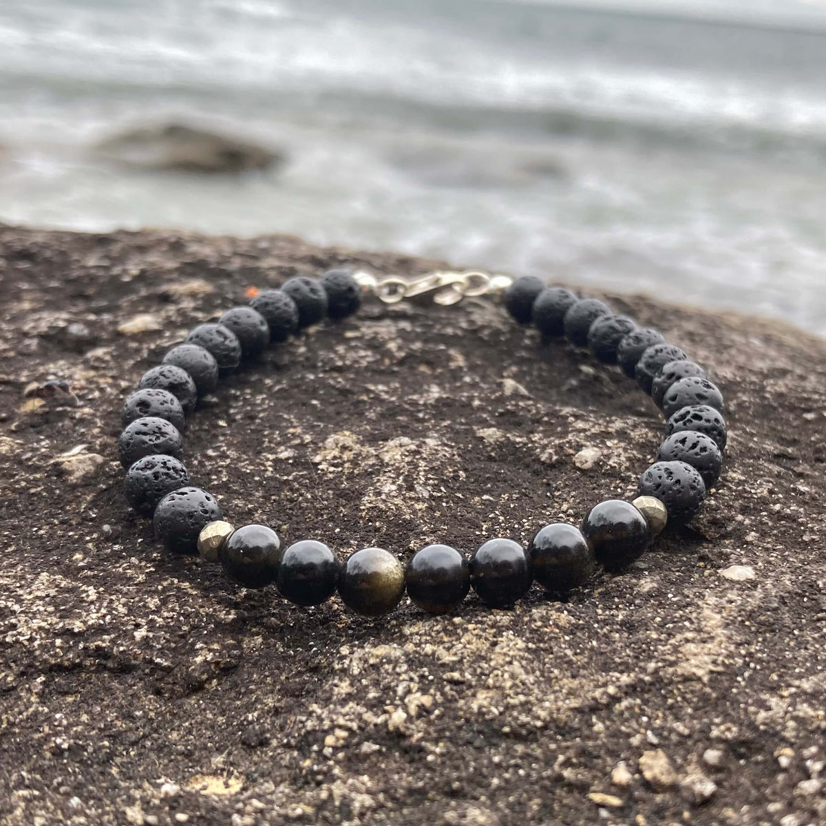 Facing Fears men's bracelet: golden obsidian with lava stone and pyrite beads, on rock by the sea