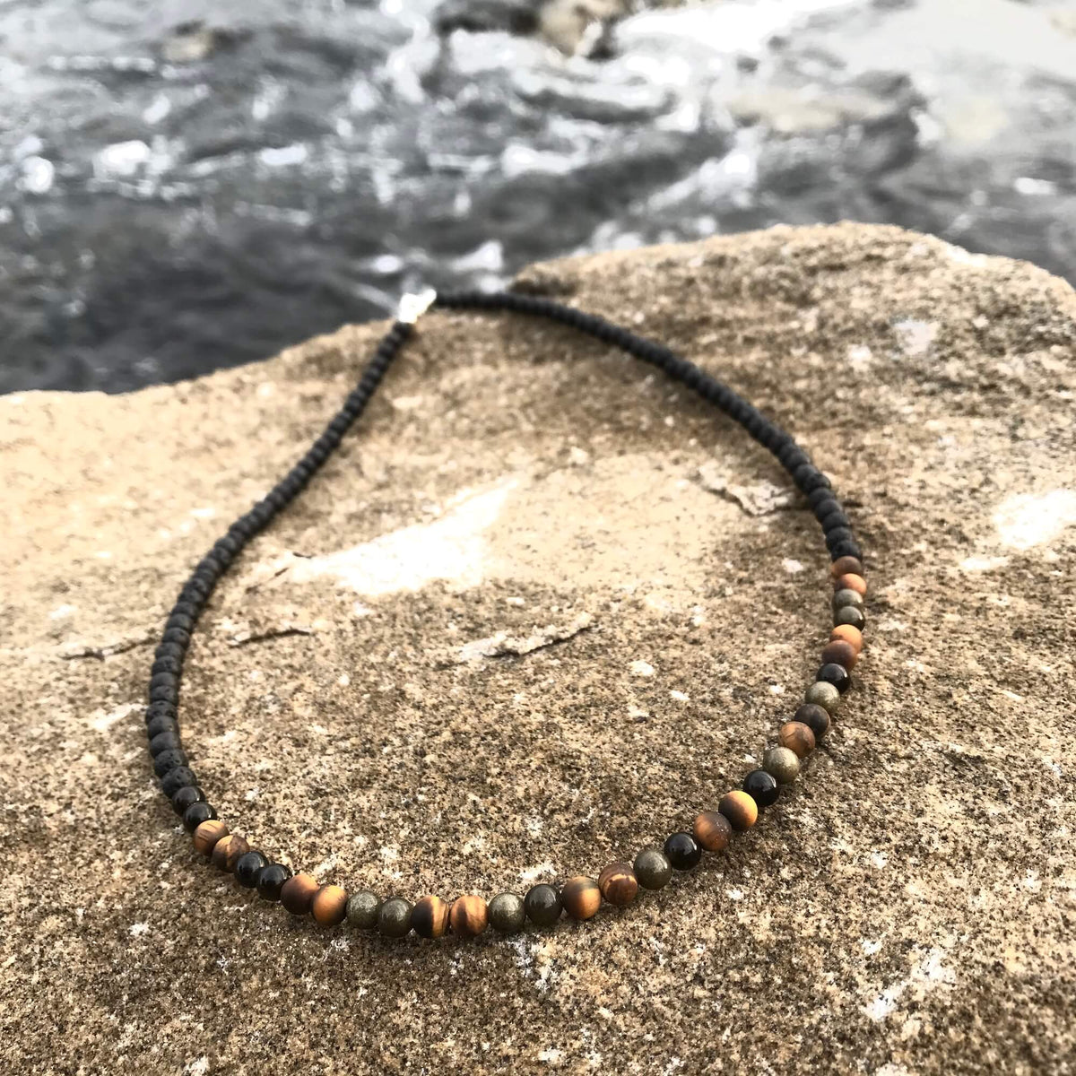 Fortitude men's necklace: golden tiger's eye and golden obsidian with black beads, on sandstone by the sea