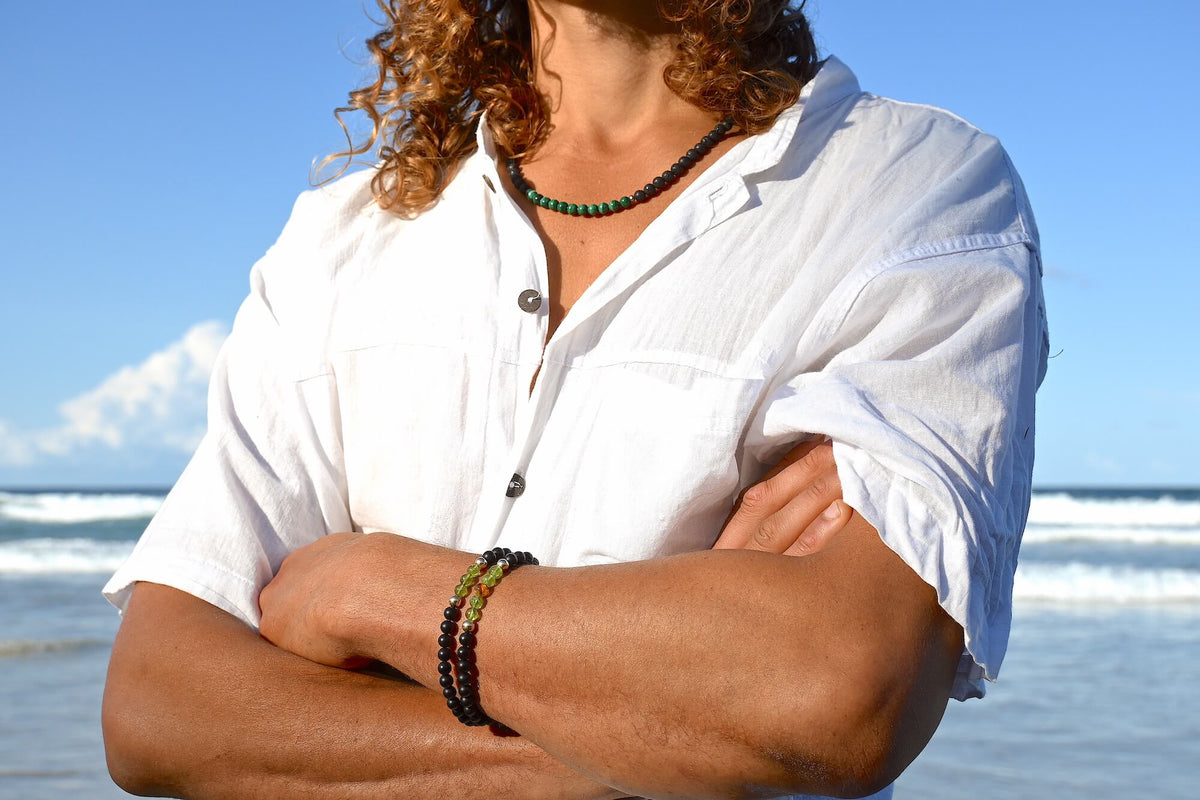 Man in a white shirt at the beach, arms crossed, wearing The Renewer bracelet