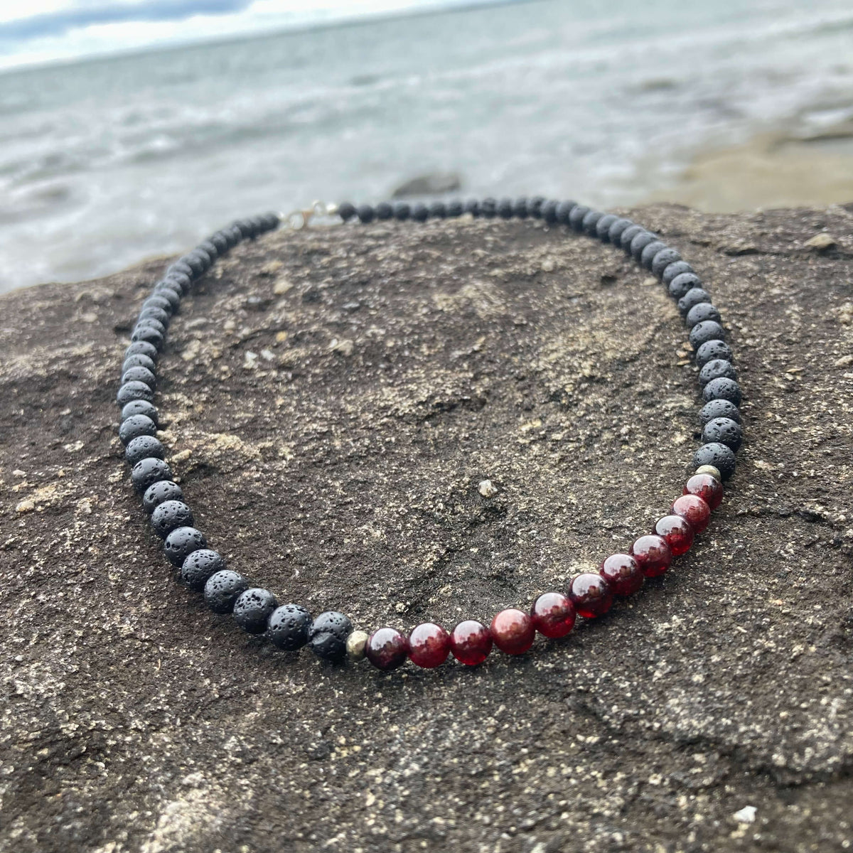 Prosperity men's necklace: deep red garnet with dark lava stone beads, laid on rock by the sea