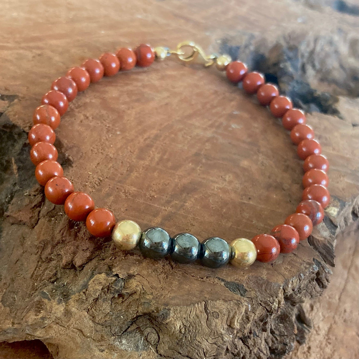 Red jasper and hematite beads on The Steadfast bracelet, resting on driftwood