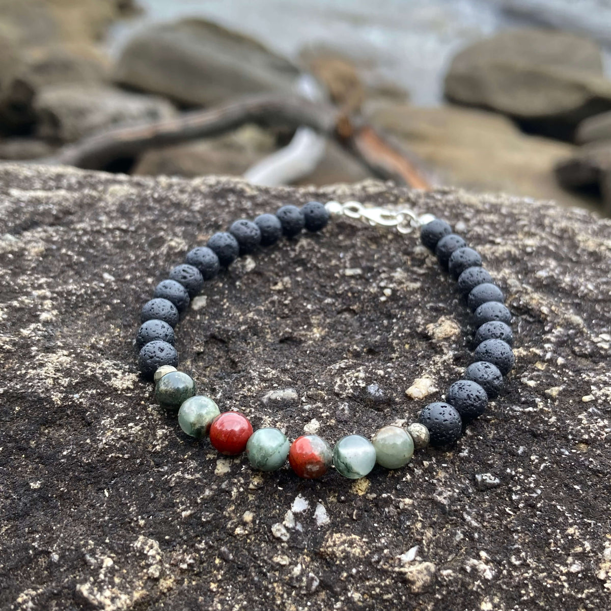 Strength men's bracelet: green-and-red bloodstone with matte black lava stone beads, on rock by the sea